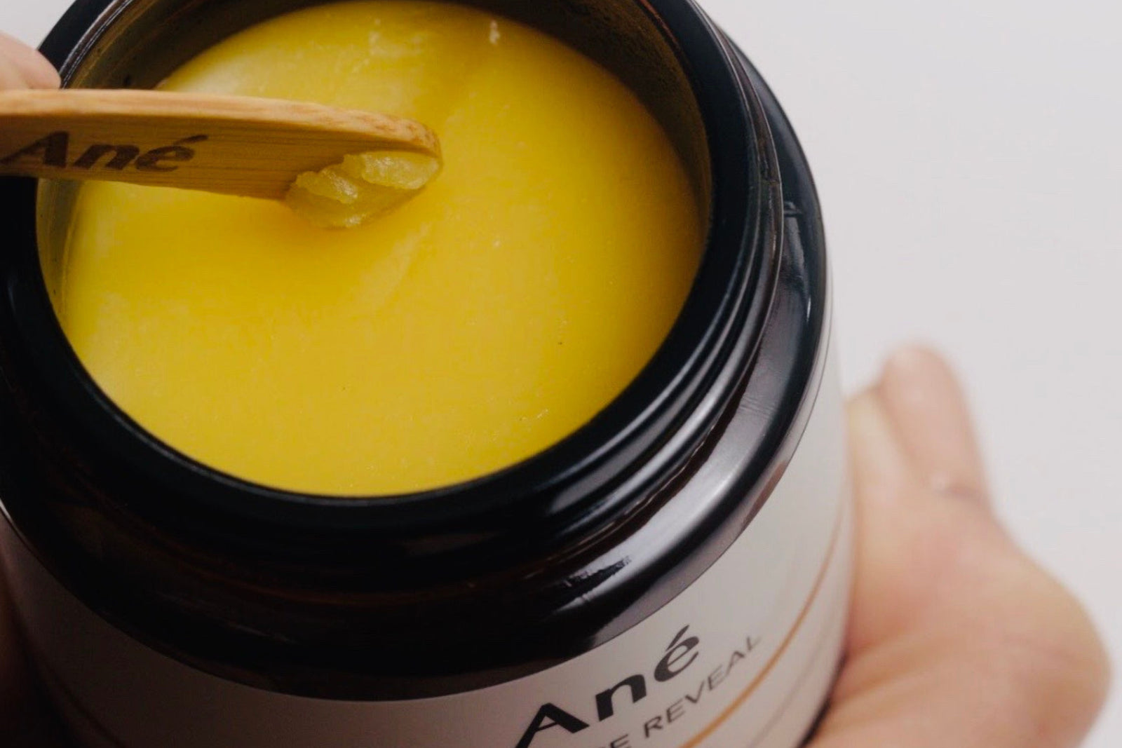 Ané Radiance Reveal Cleansing Balm sunshine yellow colour buttery soft texture being scooped out of amber glass jar with a wooded spatula 